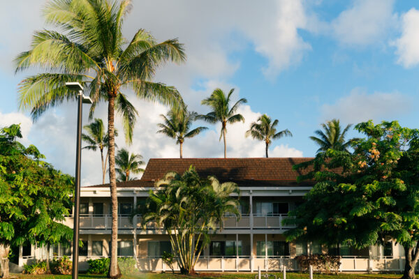 A two-story building with balconies is surrounded by lush green trees and tall palm trees under a bright blue sky with scattered clouds. Sunlight casts warm light on the scene.