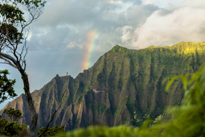 Dramatic green sea cliffs of Kauai's iconic Na Pali Coast with a brilliant rainbow arching overhead against a vivid blue sky